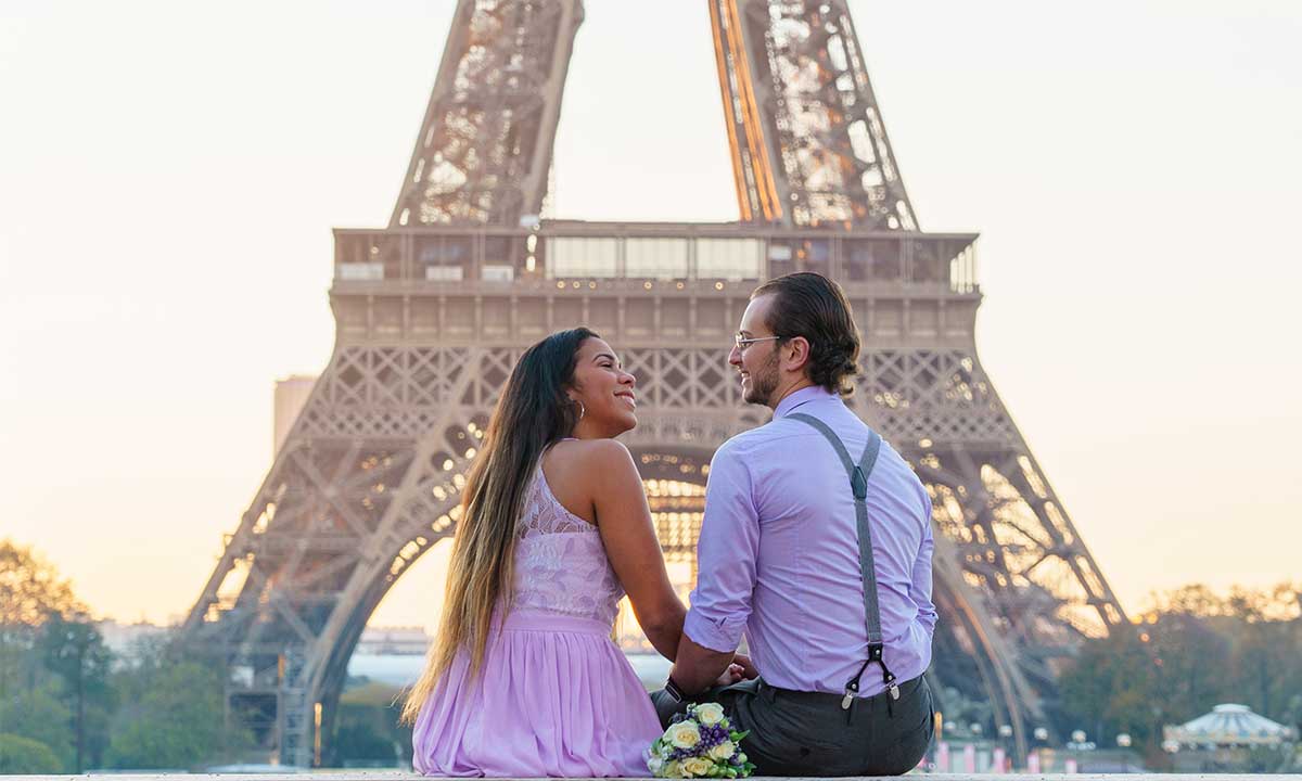 photo mariage devant la tour eiffel à Paris au lever du soleil. Mariés élégants, habillés aux couleurs mauves, le marié porte des bretelles. Le couple pose assis, de profil, en se tenant la main et se regardant