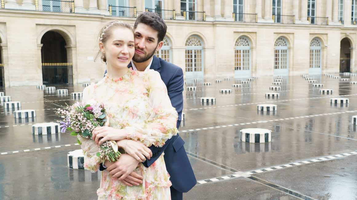photo de mariage paris Palais Royal.Au 1er plan, le couple de mariés pose face à la camera, le marié enlace la mariée, de façon romantique, les colonnes de Buren sont en arrière plan, la pluie vient de tomber. La mariée porte une robe couleur pastelle et tient un bouquet de fleurs. Le marié porte un costume bleu marine. Le couple vient de s'épouser
