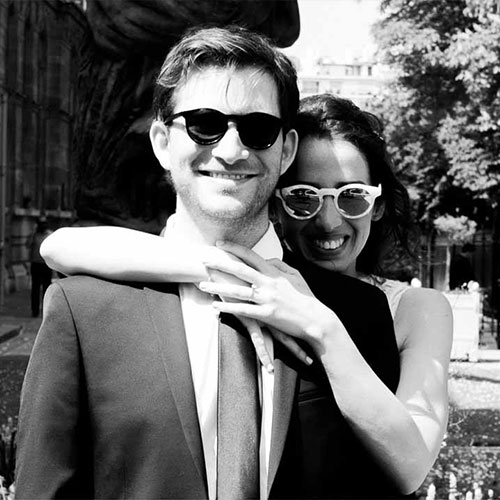 photo de mariage originale en noir et blanc. Le couple de mariés porte des lunettes de soleil. La mariée enlace le marié de façon amoureuse en posant ses mains sur son cou .